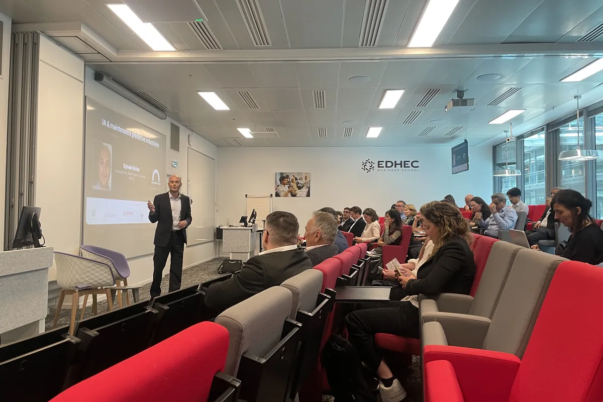 A speaker presents to a room of practitioners in an EDHEC lecture theatre with red seats